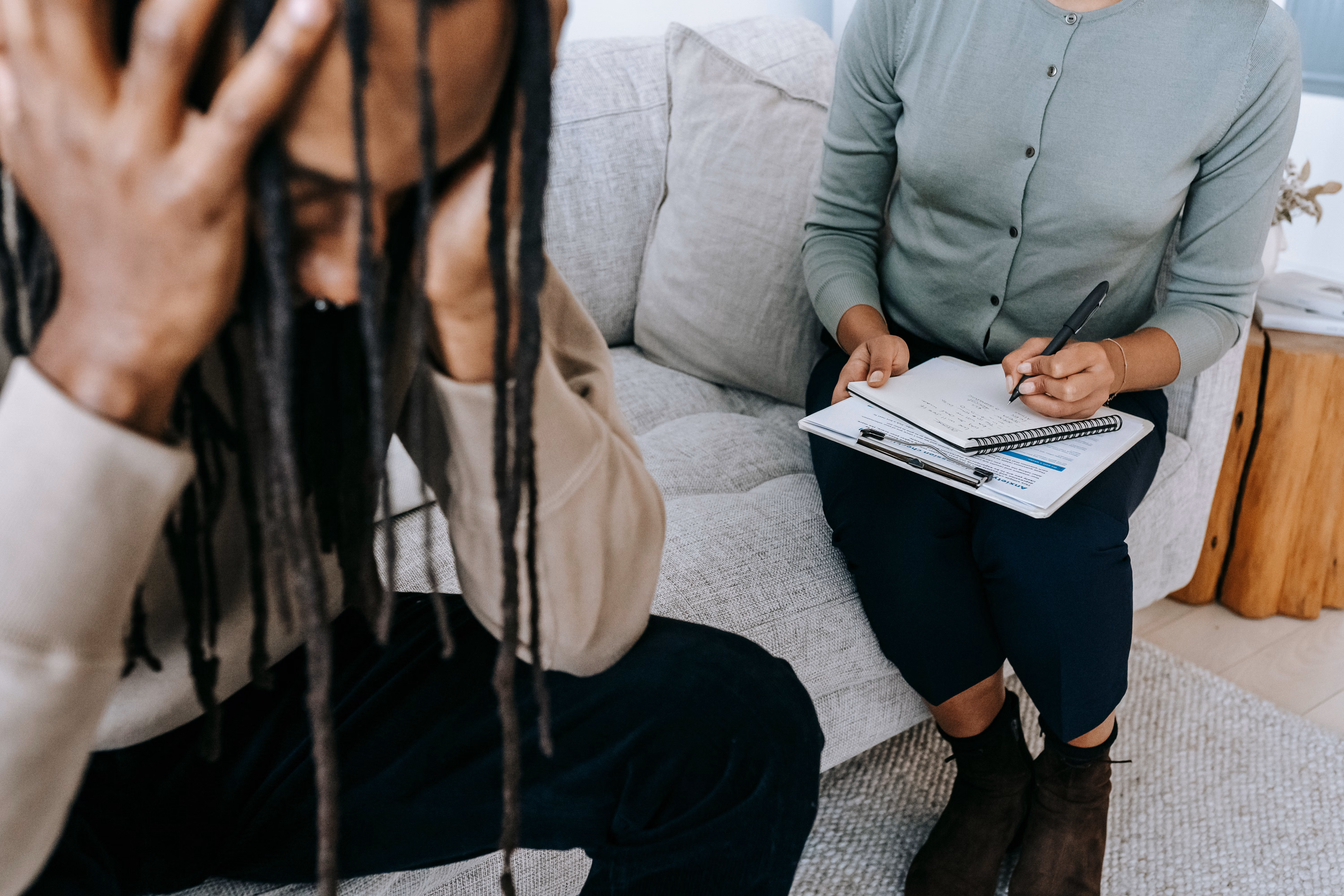 Crop concerned African American male sitting on sofa with female psychologist and holding head in hands in dismay during psychotherapy session